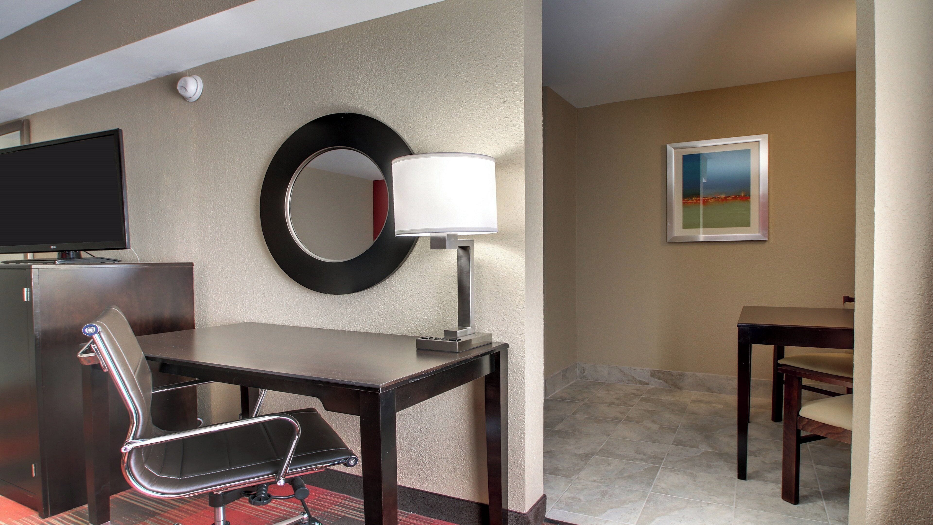 A hotel room with a desk, chair, television, lamp, mirror, and artwork on the wall.