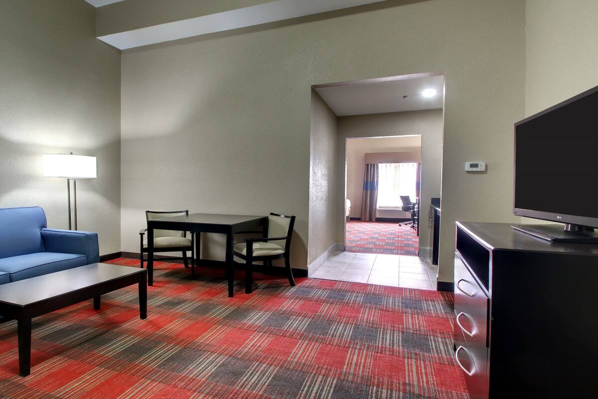 A hotel room with a blue sofa, a small table, a TV, a desk with chairs, and a carpeted floor.