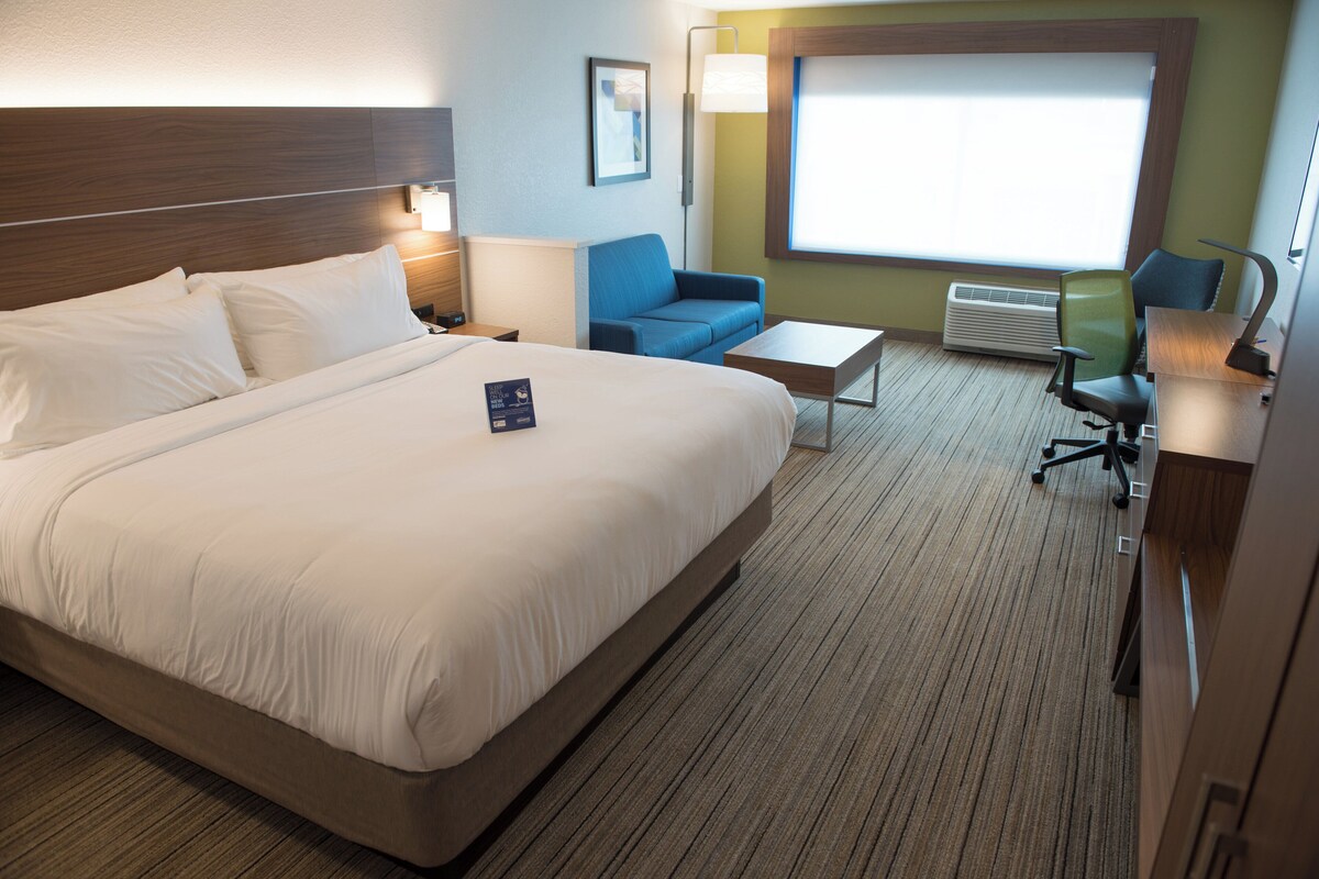 A hotel room with a large bed, a desk, a chair, a sofa, and a window with blinds.