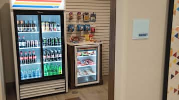A vending machine with various beverages and snacks, a small refrigerator with frozen items, and a wall sign indicating a market area.