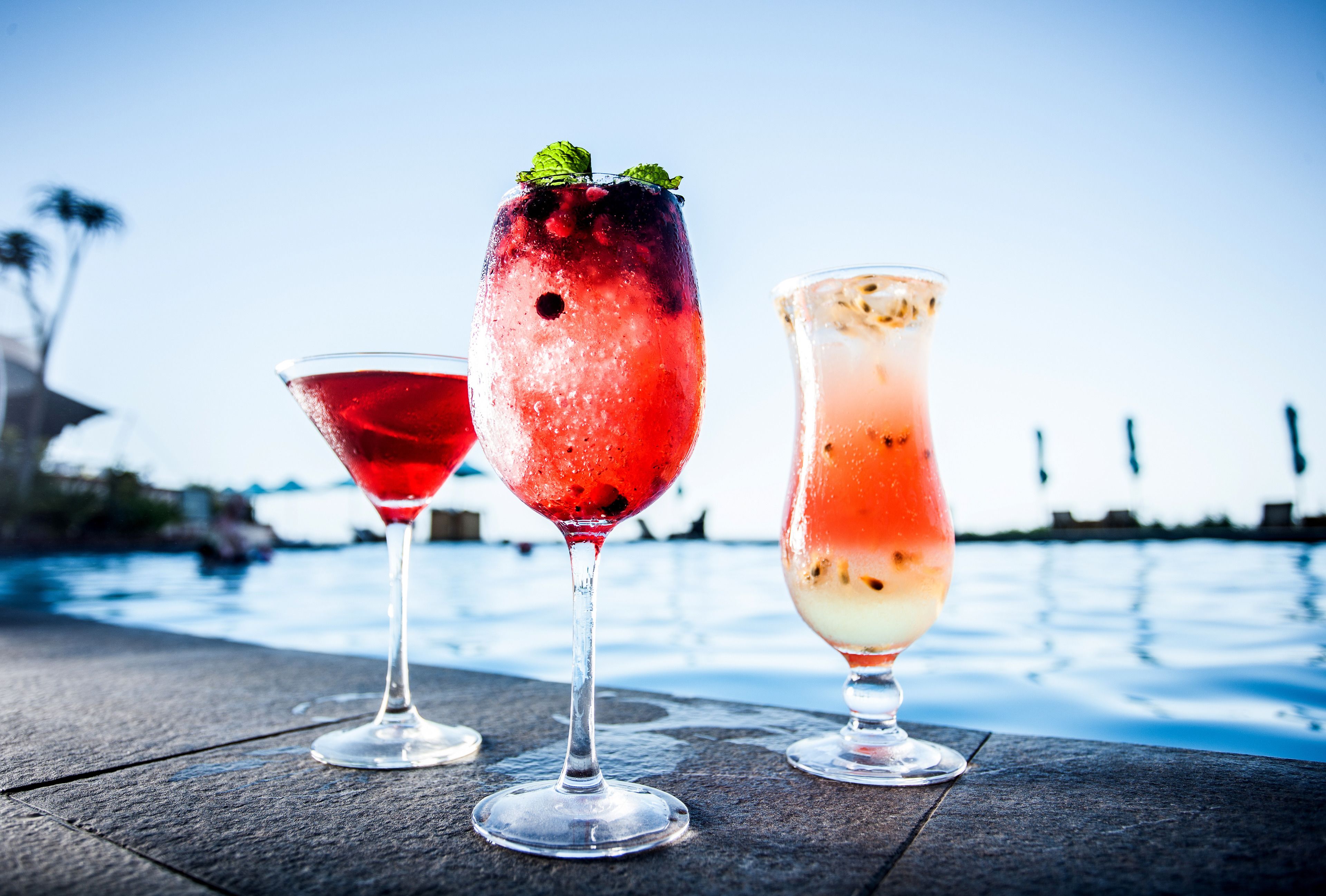 Three cocktails on a wooden table by a pool with a clear sky.