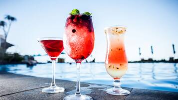 Three cocktails on a wooden table by a pool with a clear sky.