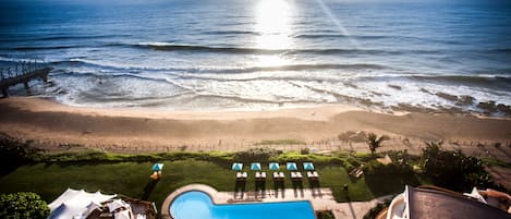 Uma área de piscina com espreguiçadeiras e guarda-sóis, vista para a praia e um pôr do sol sobre o oceano.