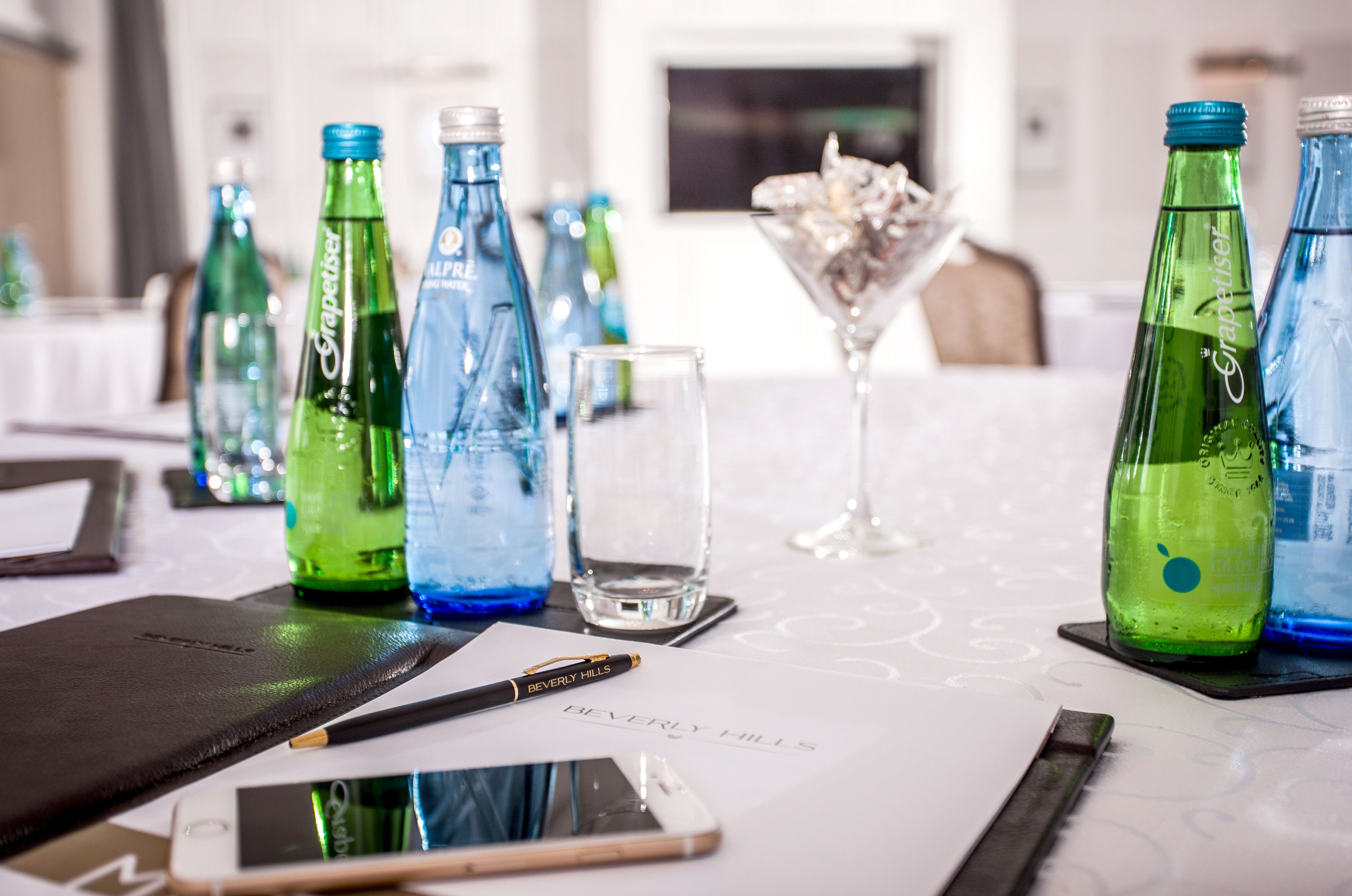 Mesa com garrafas de água, um copo de martini, um smartphone e uma caneta sobre um tecido com texto.
