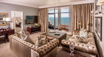 A spacious living room with a large flat-screen TV, comfortable seating, and a balcony with a view of the sea.