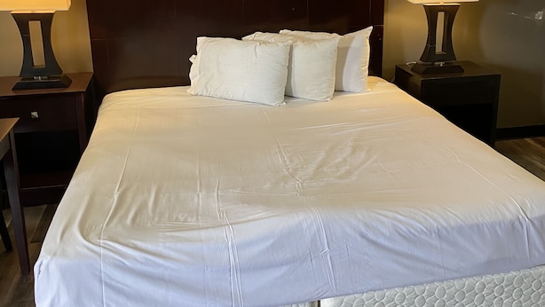 A neatly made bed with white bedding and pillows, flanked by two bedside tables with lamps, in a room with dark wooden headboard and walls. - Tourist Inn (Absecon)