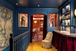 A hallway with a wooden floor, a chair, a red curtain, and a bookshelf with books and a statue. - Hotel Au Manoir Saint-Germain (Paris)