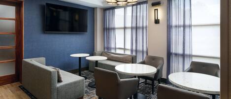 A hotel room with a flat-screen TV, a sofa, two round tables, and a large window with curtains.