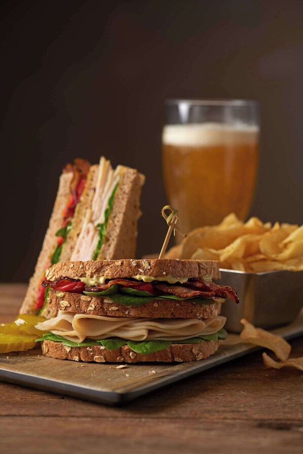 Un sándwich con un palillo, una cerveza en un vaso y un bol de papas fritas sobre una mesa de madera.
