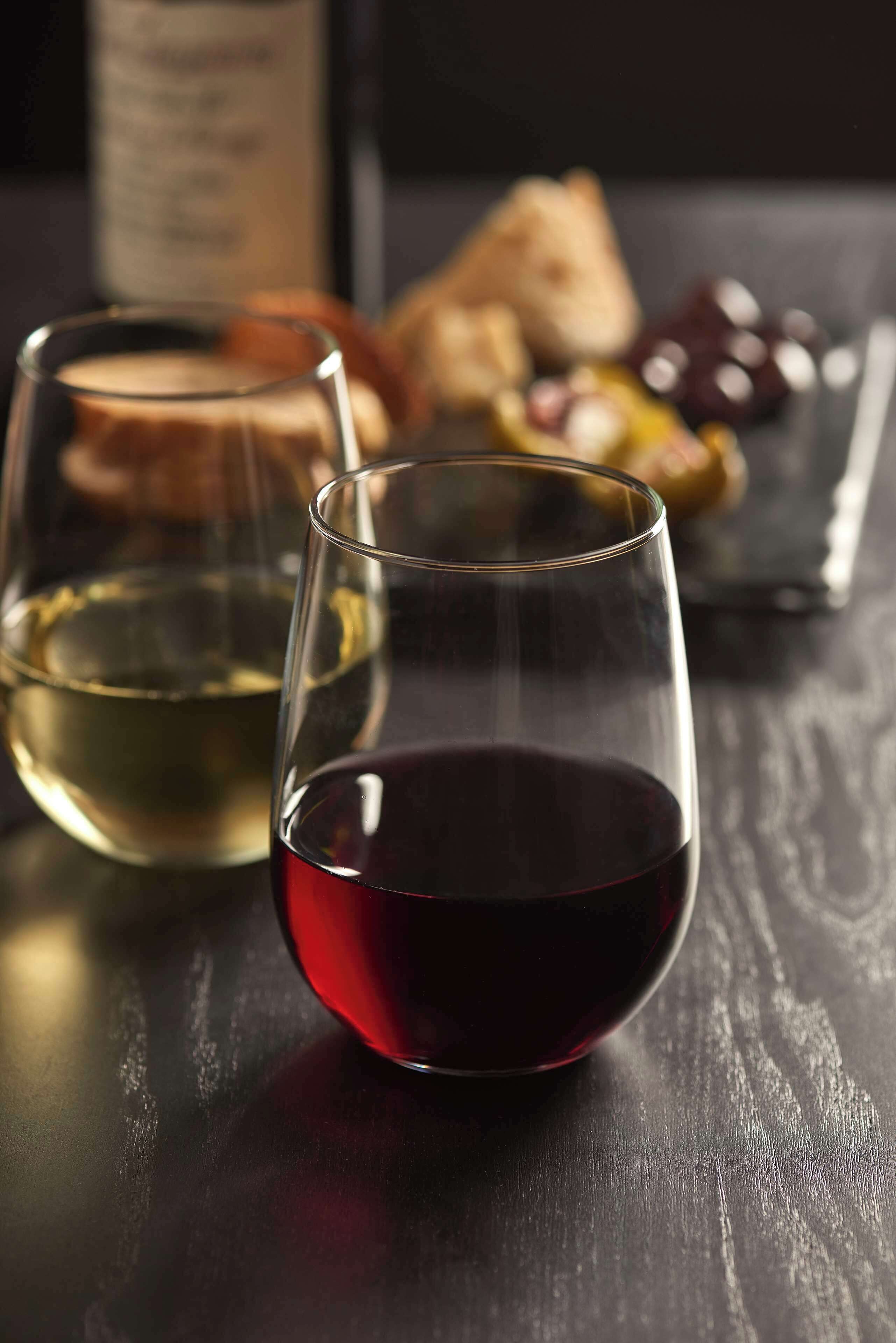 Twee glazen wijn op een houten tafel, met een fles en diverse etenswaren op de achtergrond.