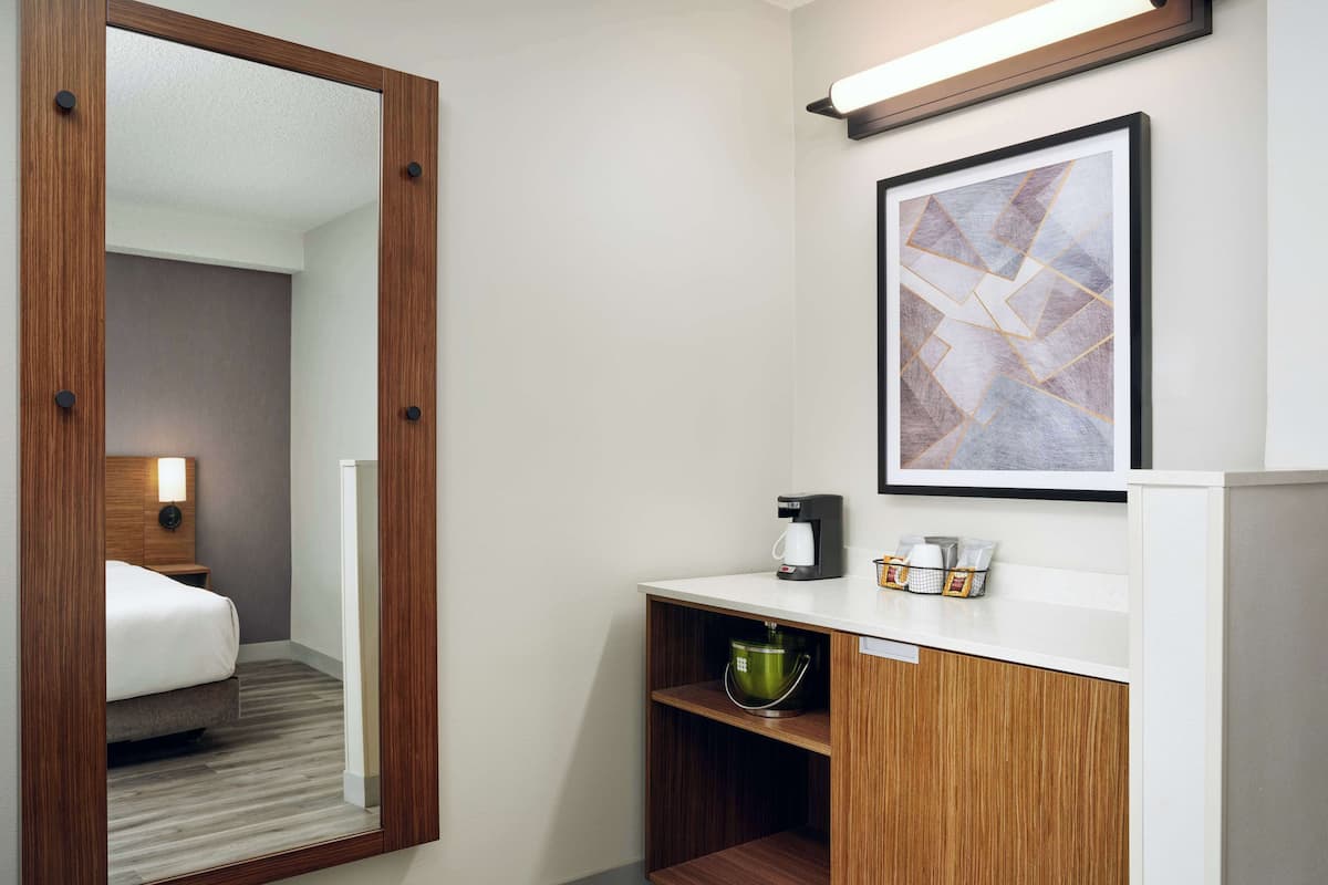 A hotel room with a bed, a mirror, a coffee maker, a bucket, and a framed picture on the wall.