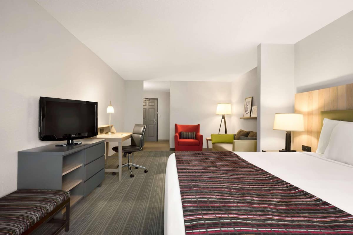 A hotel room with a bed, a desk with a chair, a television, a red chair, a green chair, and a lamp.