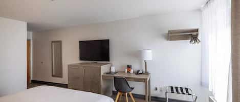 A hotel room with a bed, a desk with a chair, a television, a lamp, and a window with curtains.