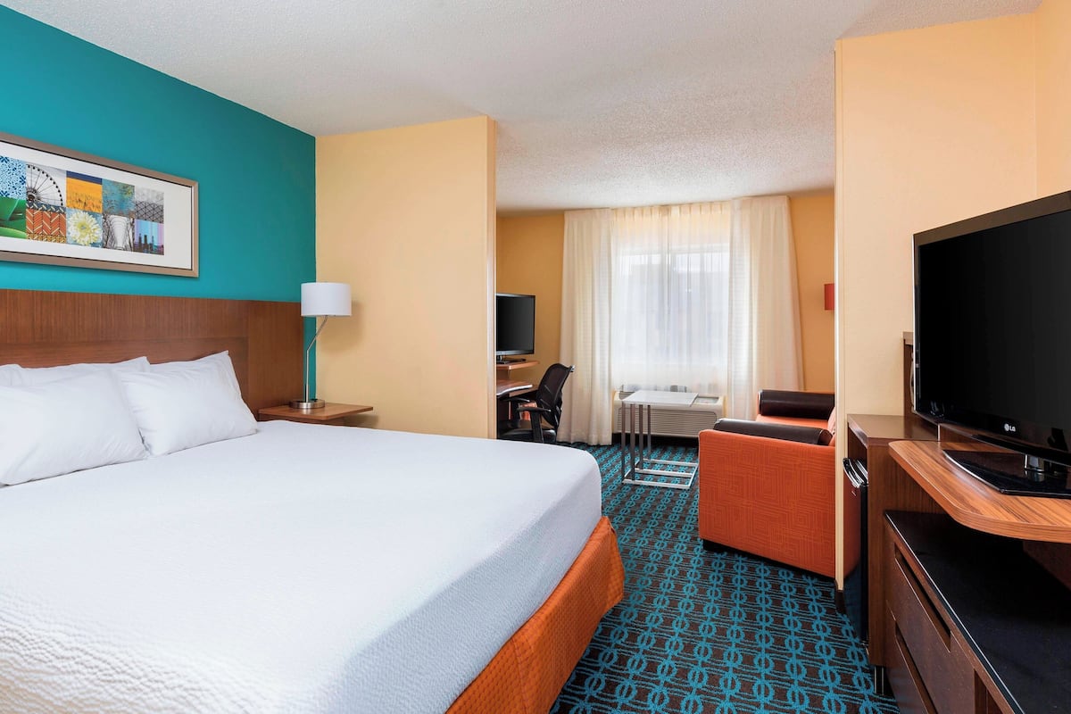 A hotel room with a large bed, a desk with a computer, a TV, and a seating area with a chair.