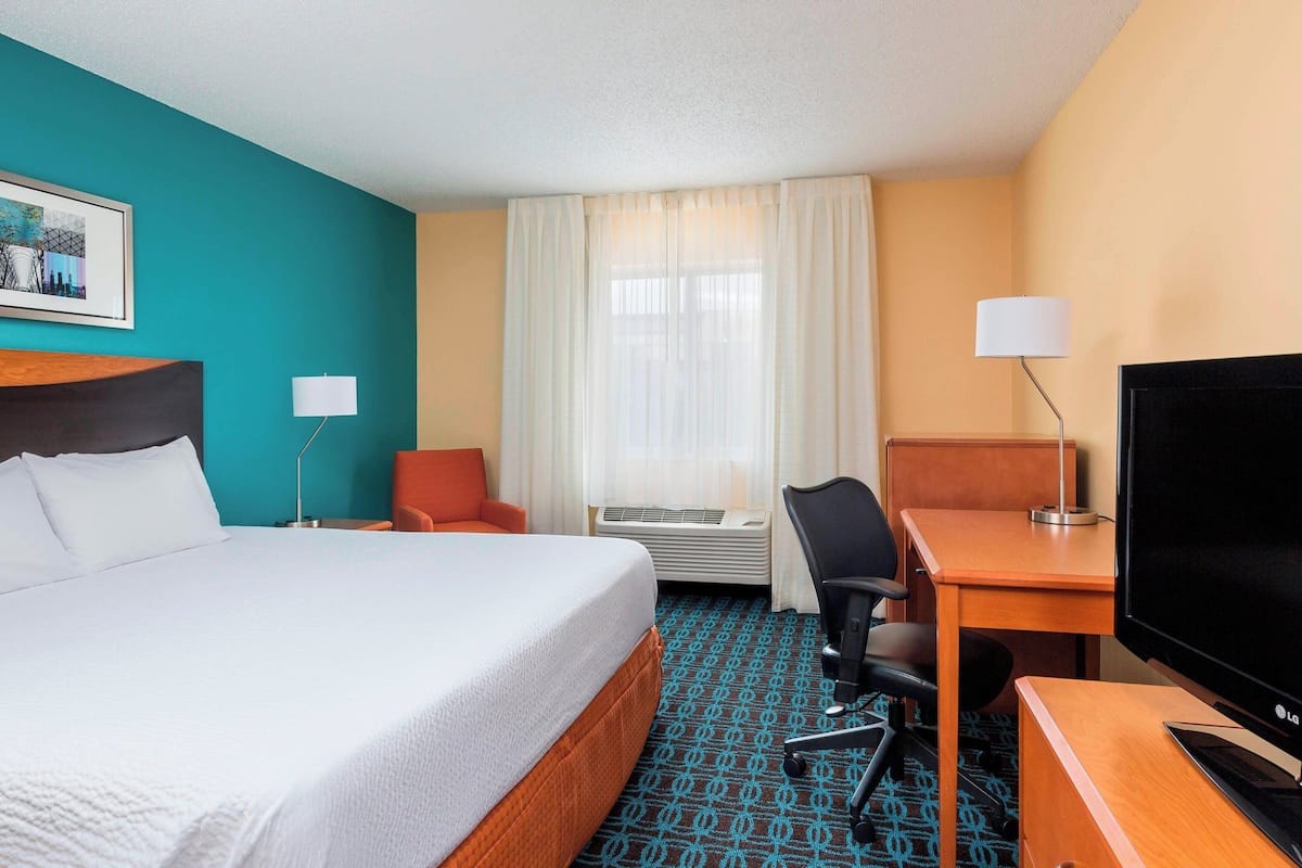 A hotel room with a large bed, a desk with a TV, a chair, a lamp, and a window with curtains.