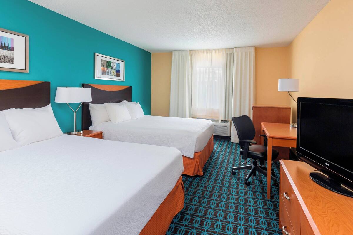A hotel room with two beds, a desk with a TV, a chair, and a window with curtains.