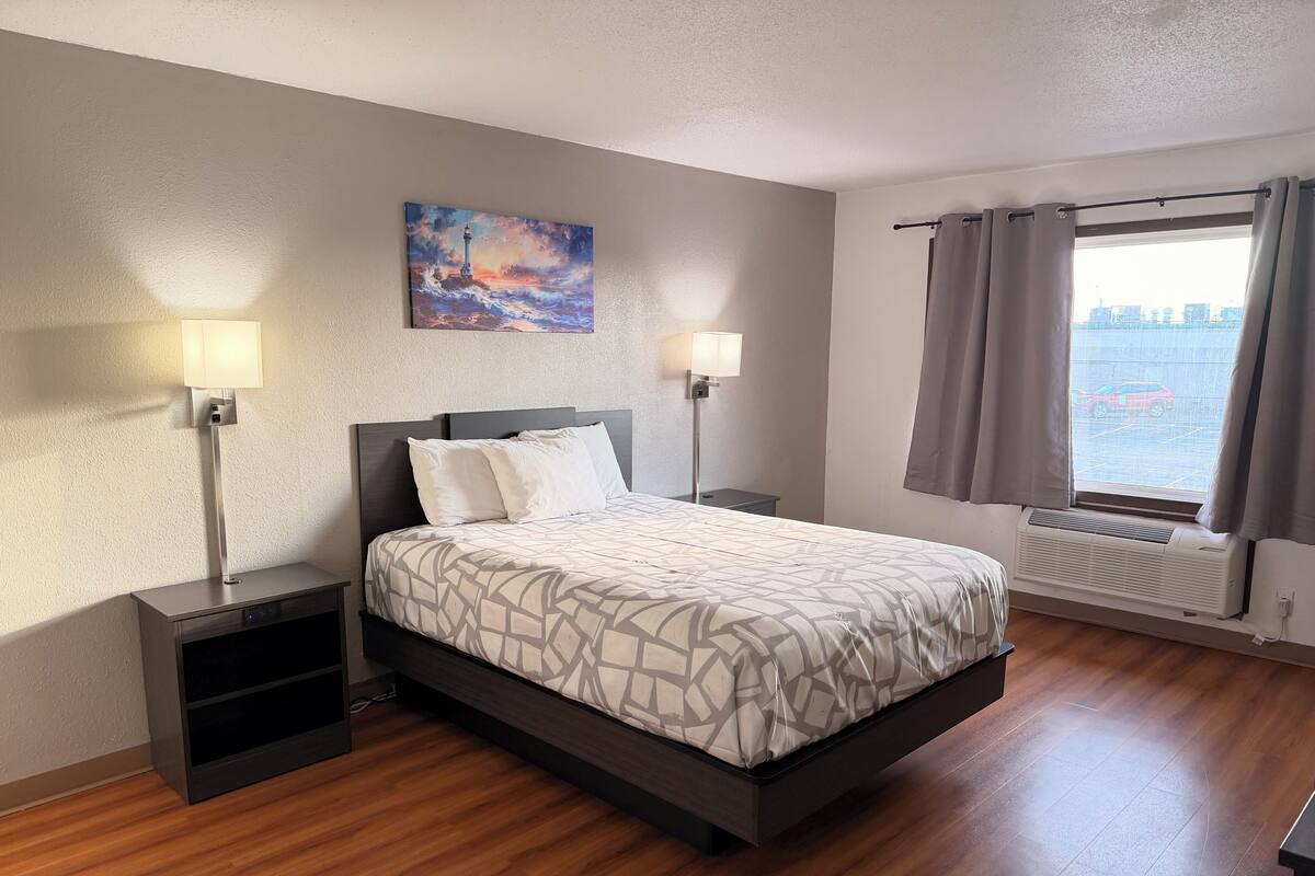 A hotel room with a bed, two bedside lamps, a painting on the wall, a window with curtains, and a radiator.