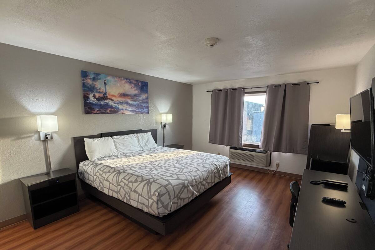 A hotel room with a bed, bedside lamps, a painting, a window with curtains, a desk with a TV, and a radiator.