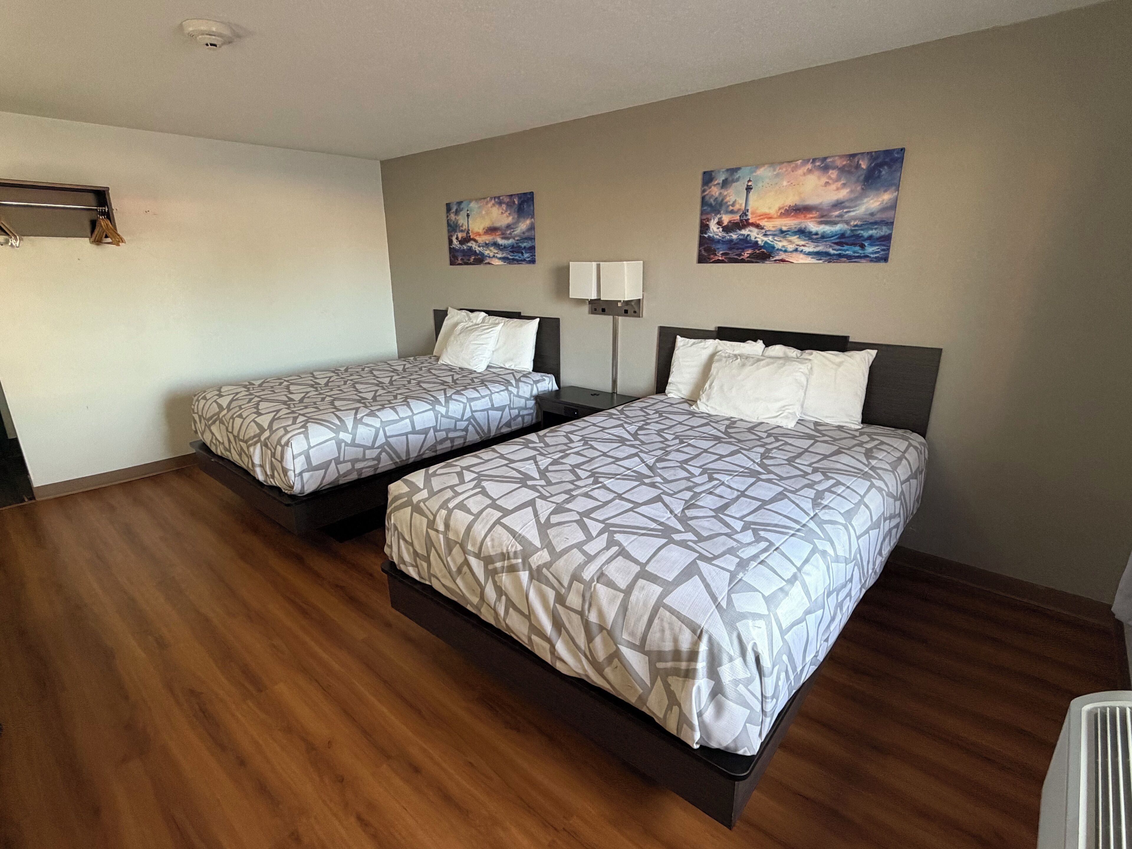 A hotel room with two beds, wooden flooring, a nightstand, a lamp, and two paintings on the wall.