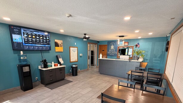 A hotel lobby with a reception desk, a large water dispenser, a trash can, a seating area with tables and chairs, and a television displaying a weather forecast.