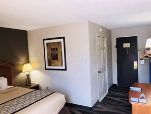 A hotel room with a bed, a framed picture on the wall, a door, and a small table with a lamp. - Days Inn by Wyndham Charlotte/Woodlawn Near Carowinds (Charlotte)