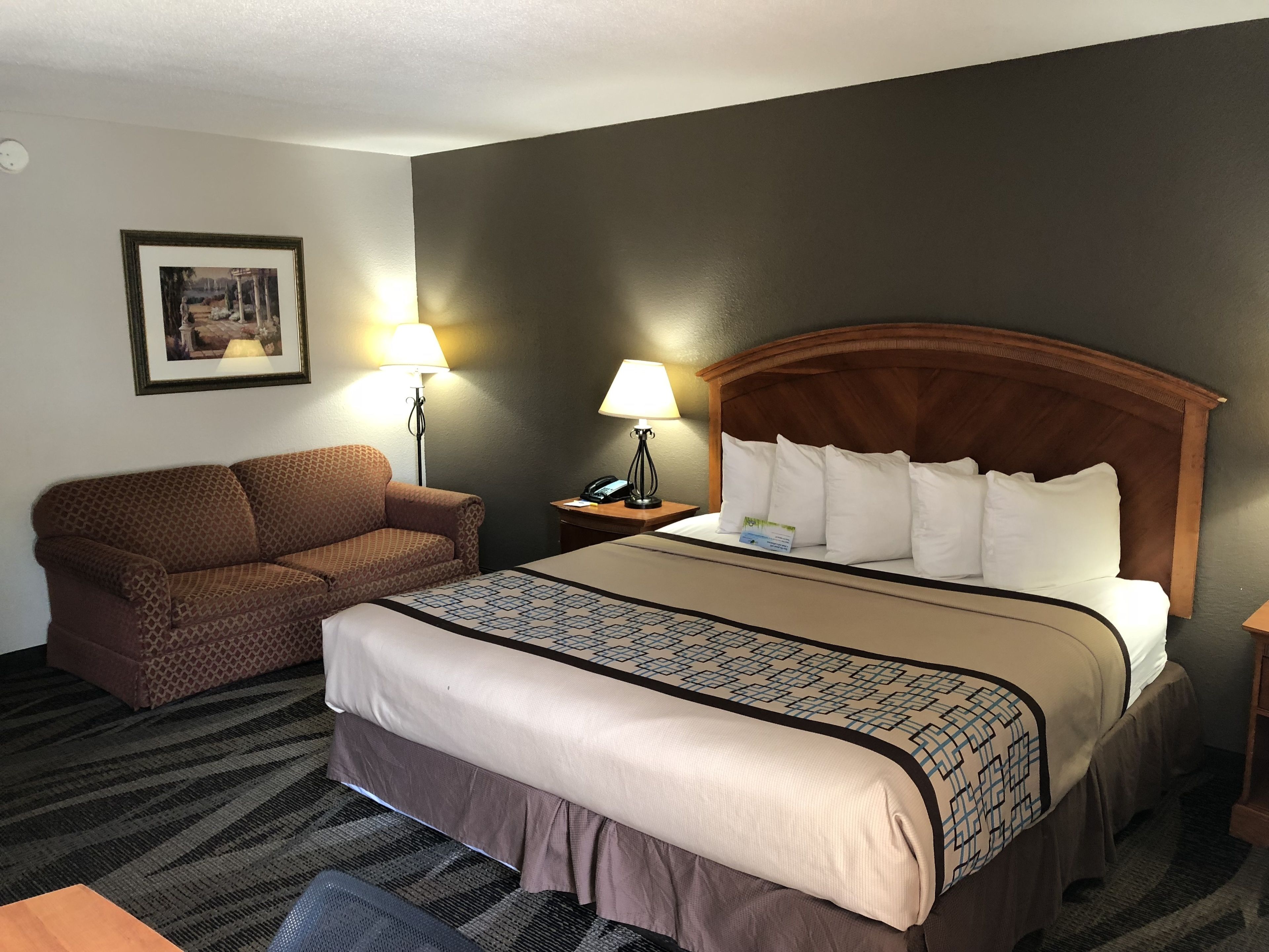 A hotel room with a bed, a sofa, two bedside lamps, a framed picture on the wall, and a carpeted floor.
