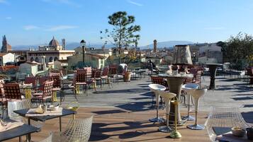 Un espacio de comedor al aire libre con mesas y sillas, una fuente y vistas de la ciudad al fondo.