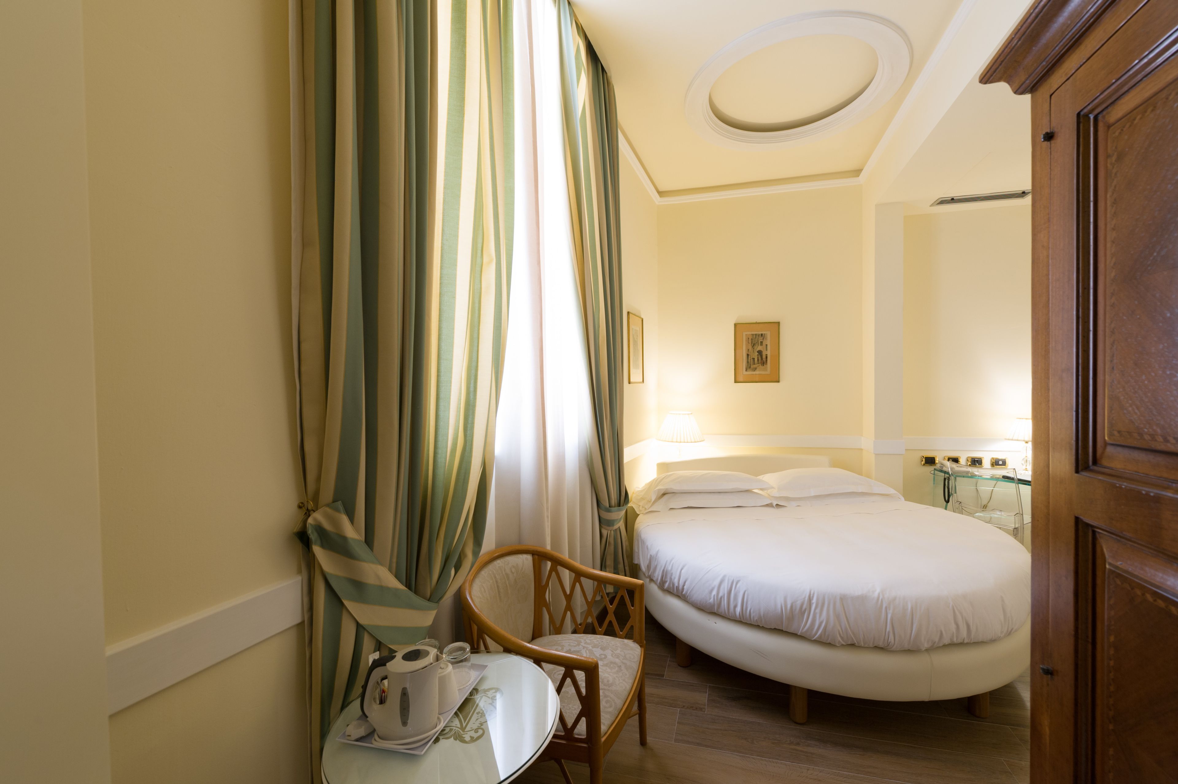 A hotel room with a bed, a chair, a table with a laptop and a cup, and a window with curtains.