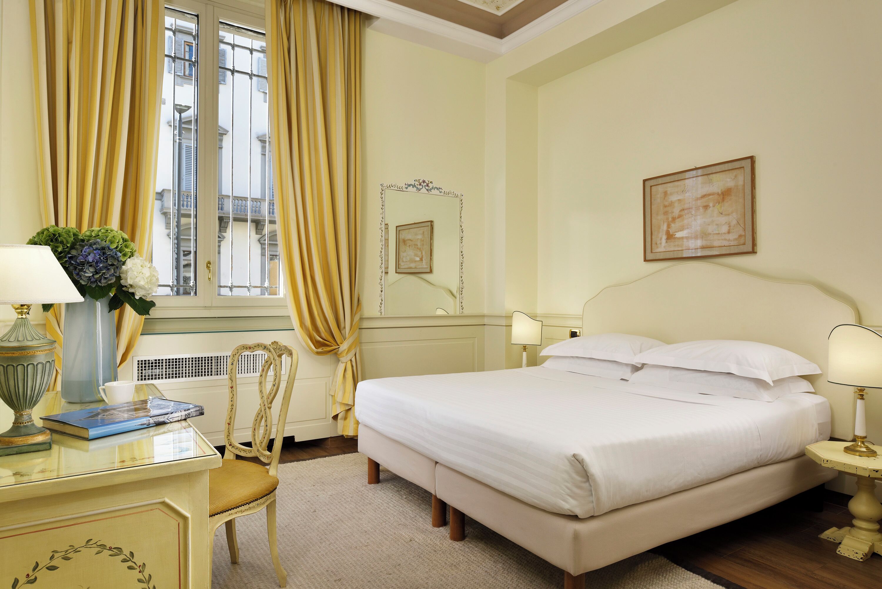 A hotel room with a bed, a desk with a lamp, a chair, a window with curtains, and a framed picture on the wall.