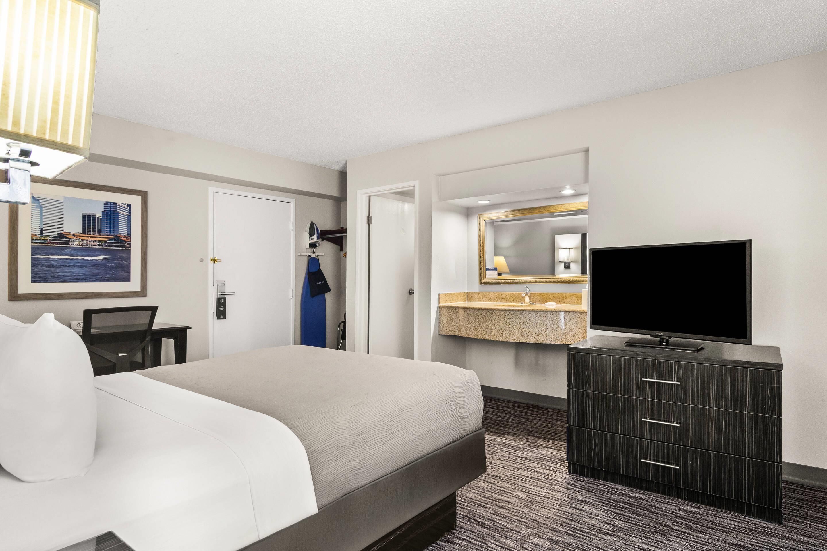 Habitación de hotel con una cama, televisor, cómoda, baño con lavamanos y un cuadro en la pared.