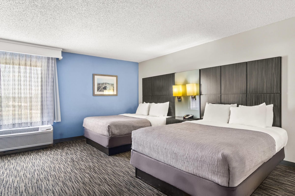 A hotel room with two beds, a large window with curtains, a wall-mounted lamp, and a framed picture.