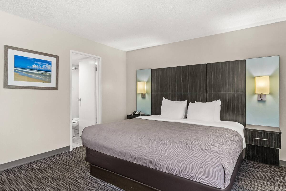 A hotel room with a large bed, two bedside lamps, a headboard, a nightstand, a framed picture on the wall, and a door leading to a bathroom.