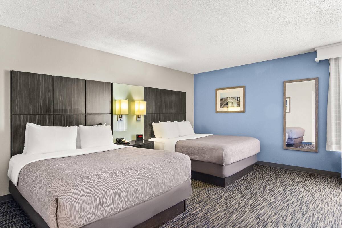 A hotel room with two beds, a large mirror, a nightstand, and a wall-mounted lamp.