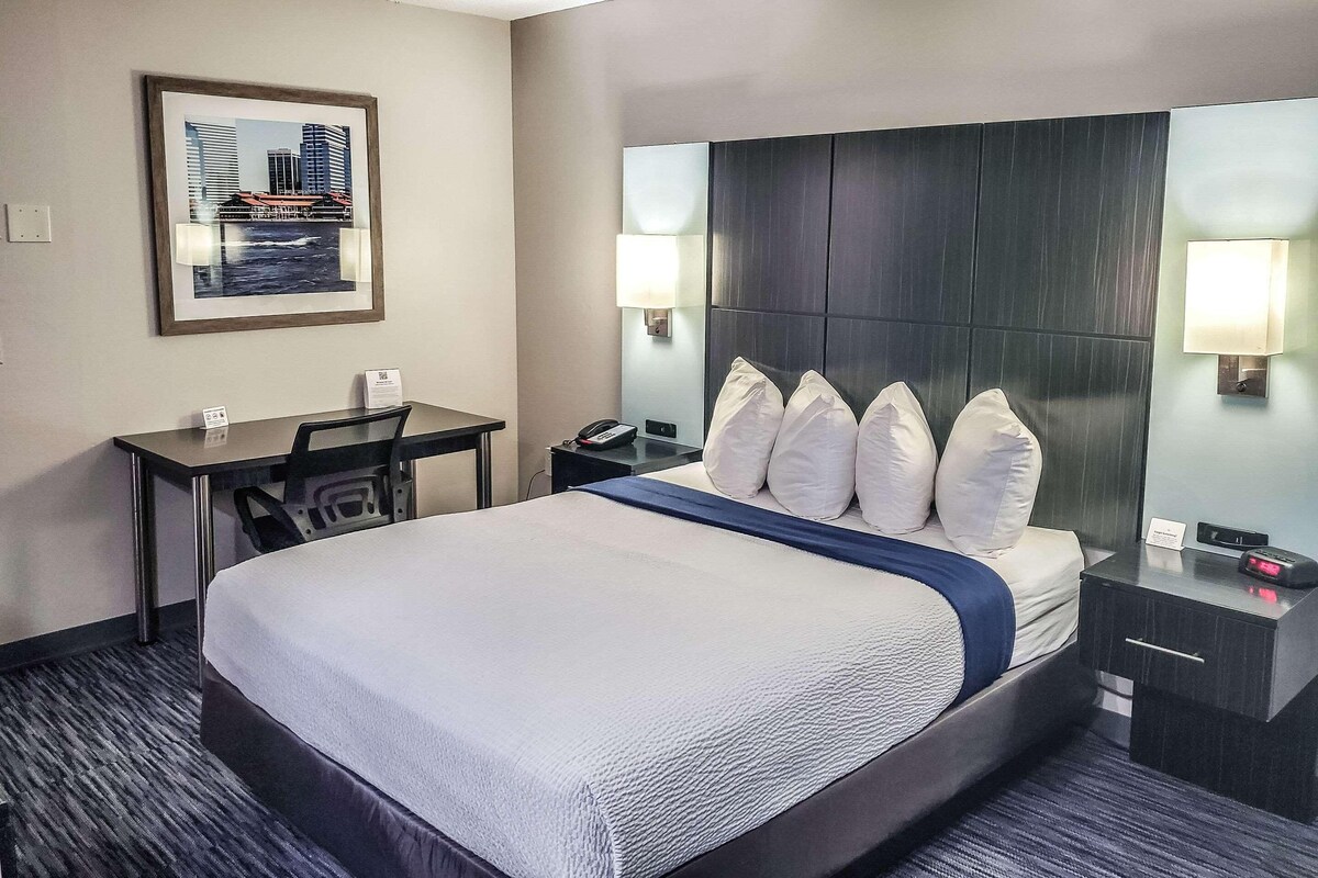 A hotel room with a large bed, a desk with a chair, a phone, and a nightstand with a lamp.