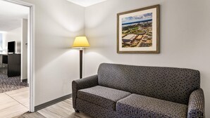 Habitación de hotel con un sofá estampado, un cuadro enmarcado de una vista urbana, una lámpara de pie y una puerta que da a otra habitación.