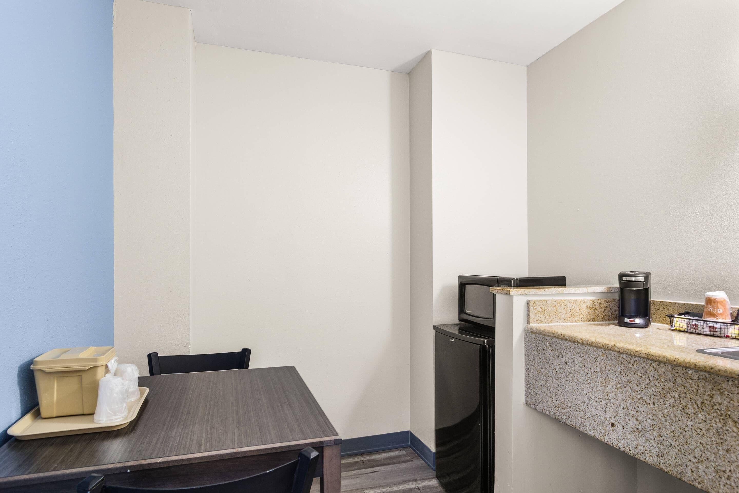 A small kitchenette with a microwave, coffee maker, and a small refrigerator. There is a table with chairs and a tray with a container and a plastic bag.