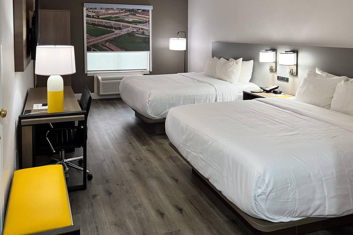A hotel room with two beds, a desk with a lamp, a chair, and a view of an airport.