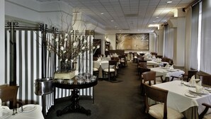 Un comedor formal con varias mesas dispuestas para el servicio, un jarrón decorativo con ramas y una partición a rayas.
