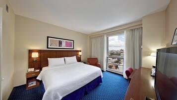 A hotel room with a large bed, a desk with a TV, a chair, a window with curtains, and a framed picture on the wall.