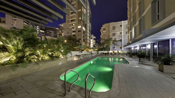 Zona de piscina nocturna con piscina verde, palmeras y edificios modernos al fondo.