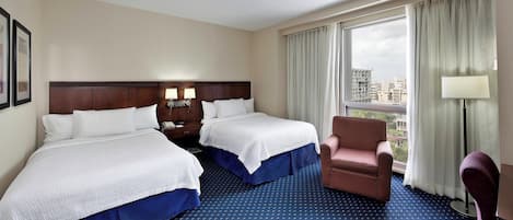 Ein Hotelzimmer mit zwei Betten, einem Sessel, einem Schreibtisch und einem großen Fenster mit Vorhängen.