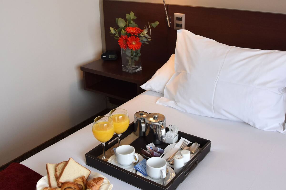 Ein ordentlich bezogenes Bett mit weißen Leinen, ein Tablett mit Frühstücksartikeln, darunter Croissants, Toast, Obst und Kaffee, sowie ein Nachttisch mit Lampe und einer Blumenvase.