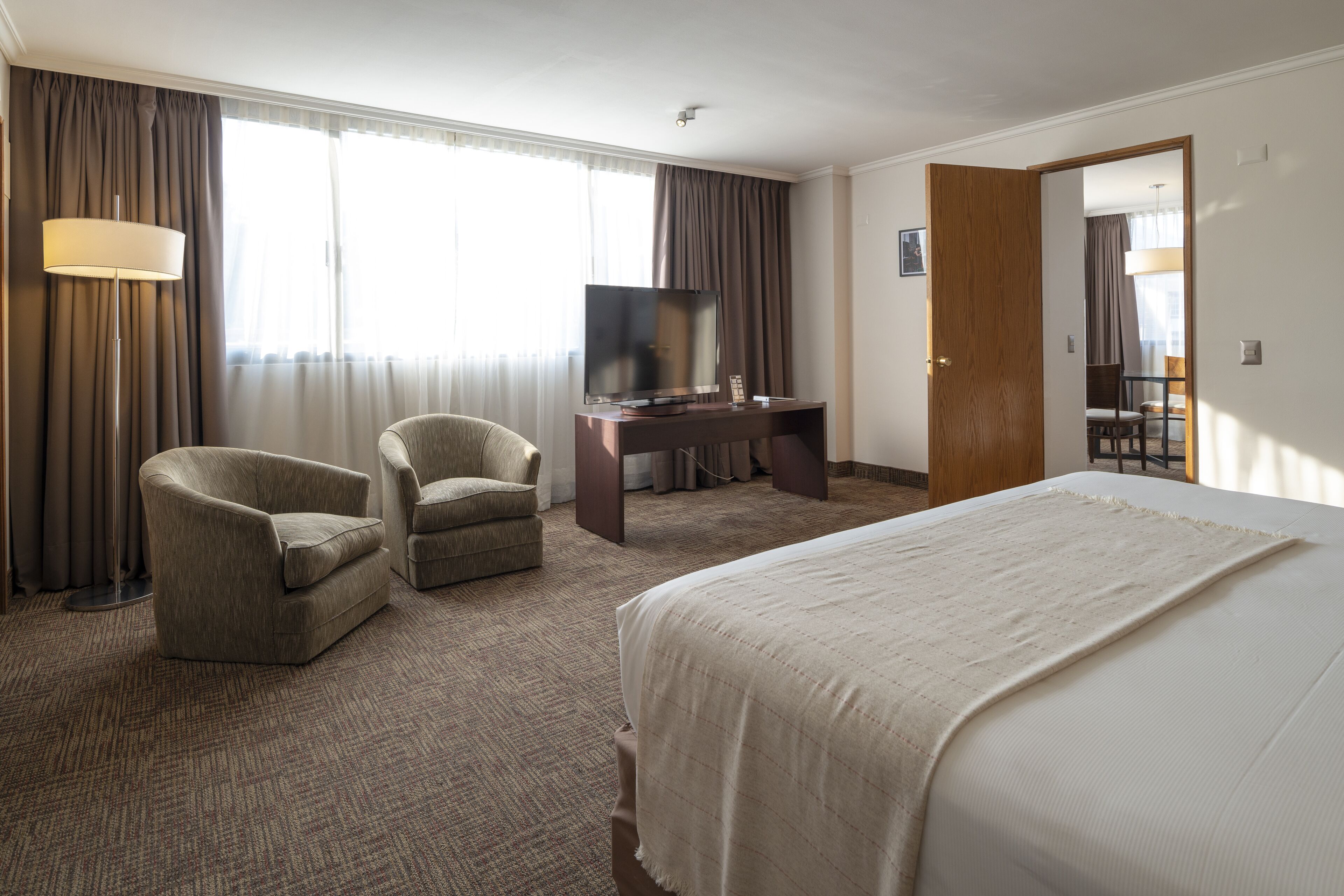 A hotel room with a bed, two armchairs, a desk with a TV, and a large window with curtains.