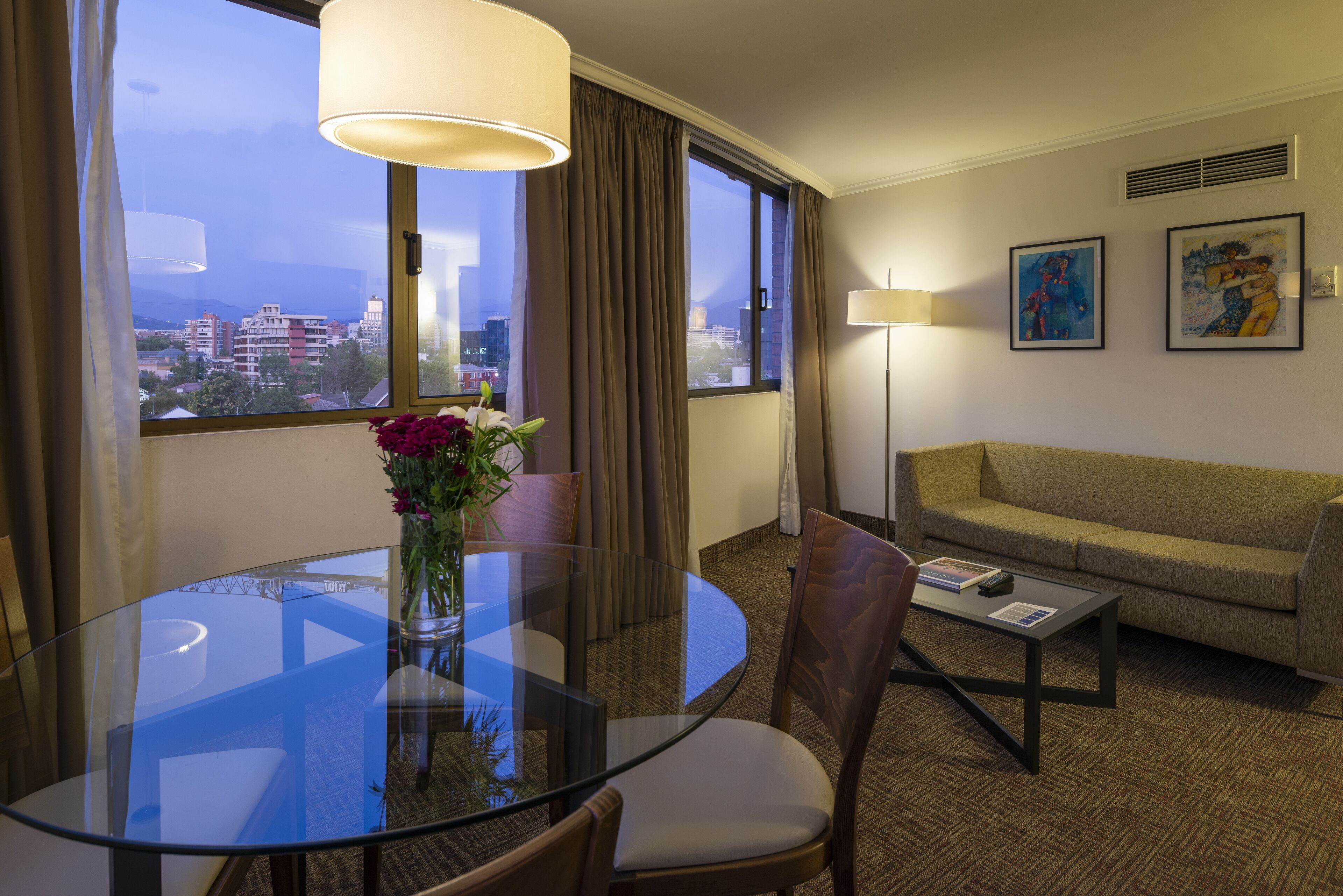 A hotel room with a glass dining table, a sofa, a coffee table, and a view of the cityscape.