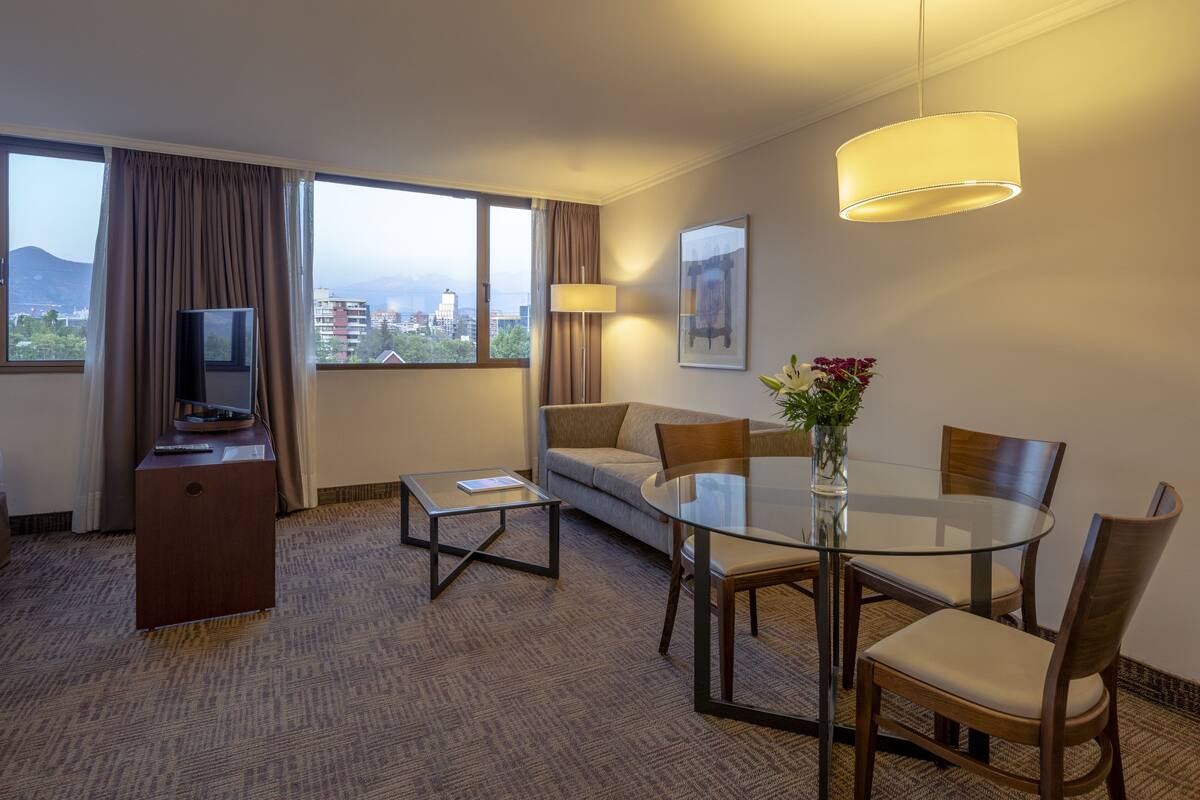 Ein Hotelzimmer mit einer Couch, einem Glastisch, einem Fernseher auf einer Holzkommode und einem großen Fenster mit Vorhängen.
