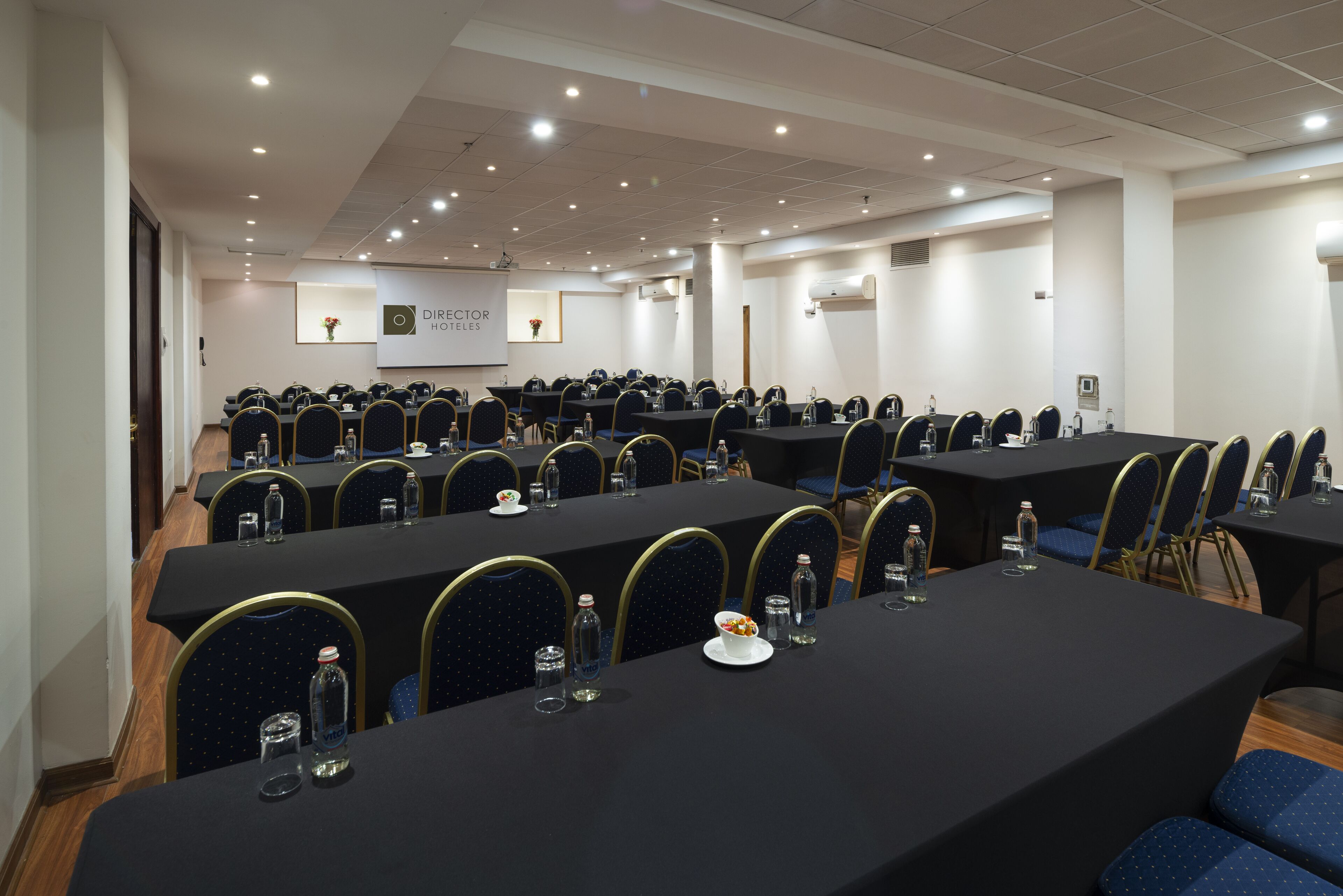 Un salón de conferencias con varias mesas y sillas, equipado con botellas de agua y una pantalla de proyector.