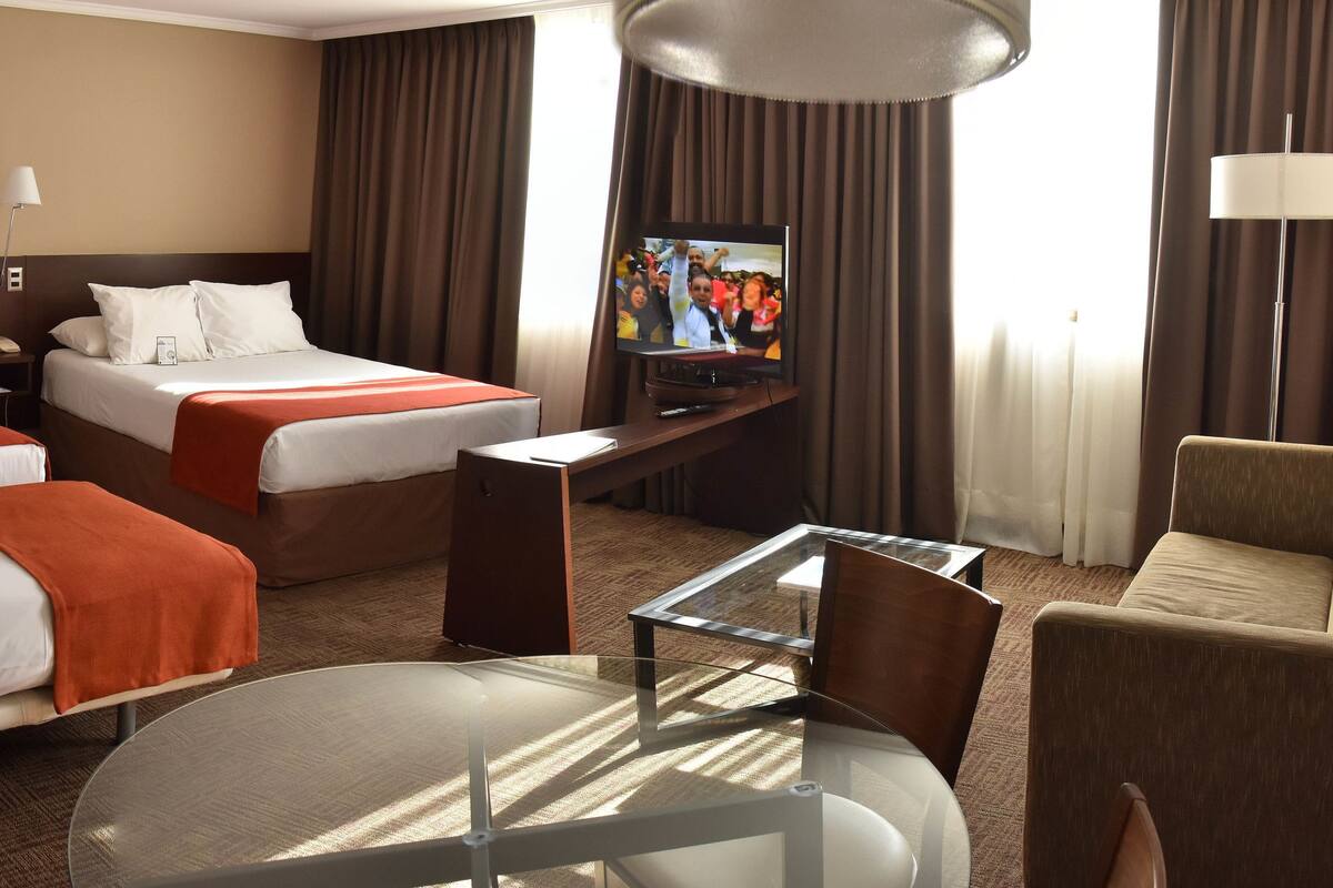 Ein Hotelzimmer mit zwei Betten, einem Fernseher, einem Glastisch und einem Sofa.