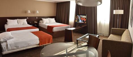 Quarto de hotel com duas camas, uma televisão, uma mesa de vidro e um sofá.