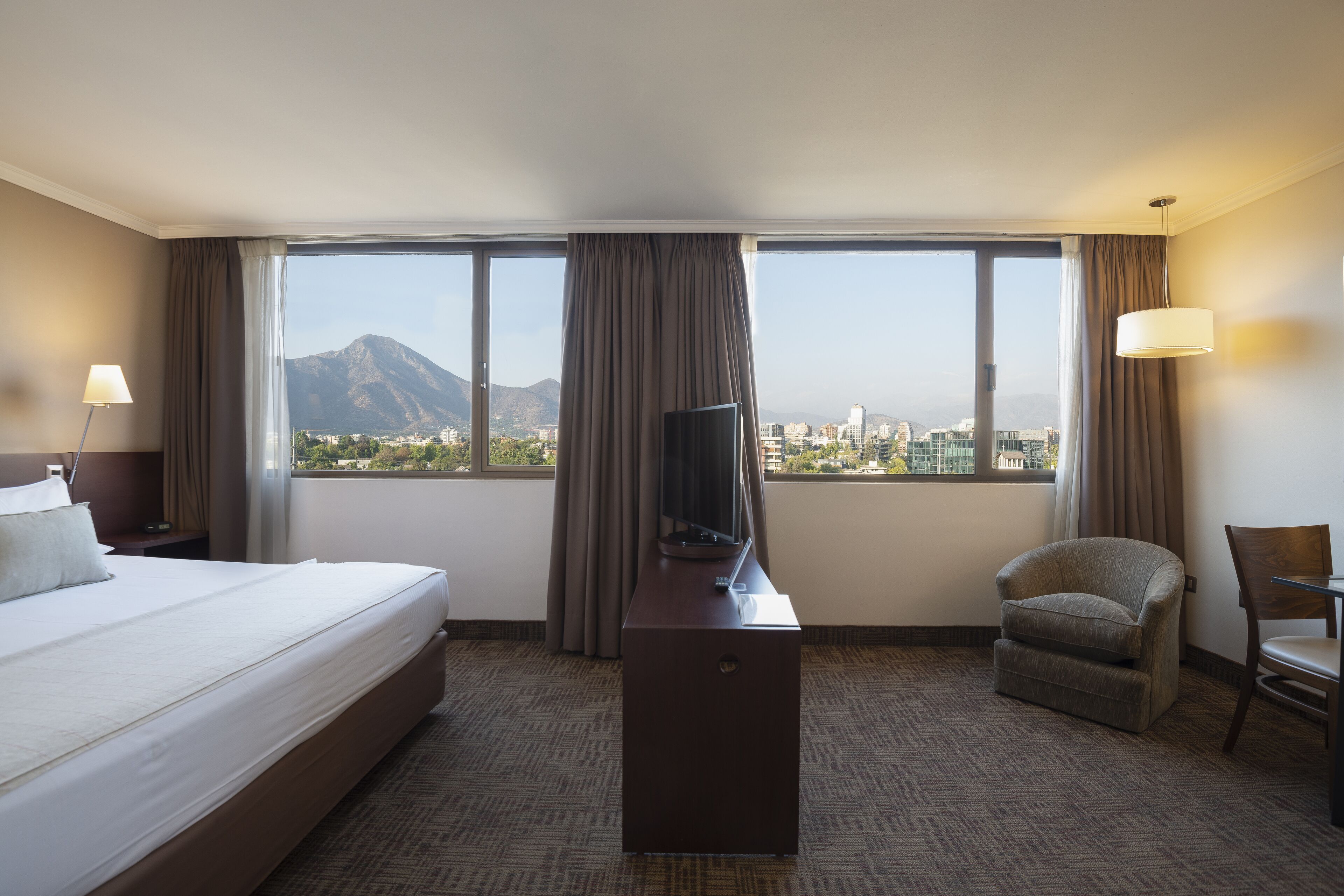 A hotel room with a large bed, a desk with a television, a chair, and a view of a cityscape and mountains.