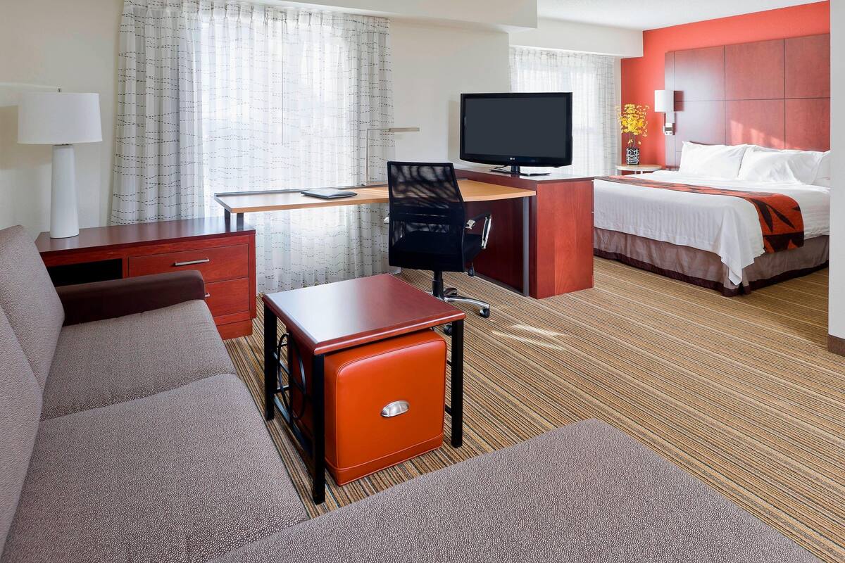 A hotel room with a bed, a desk with a computer, a TV, a sofa, and a small orange cabinet.
